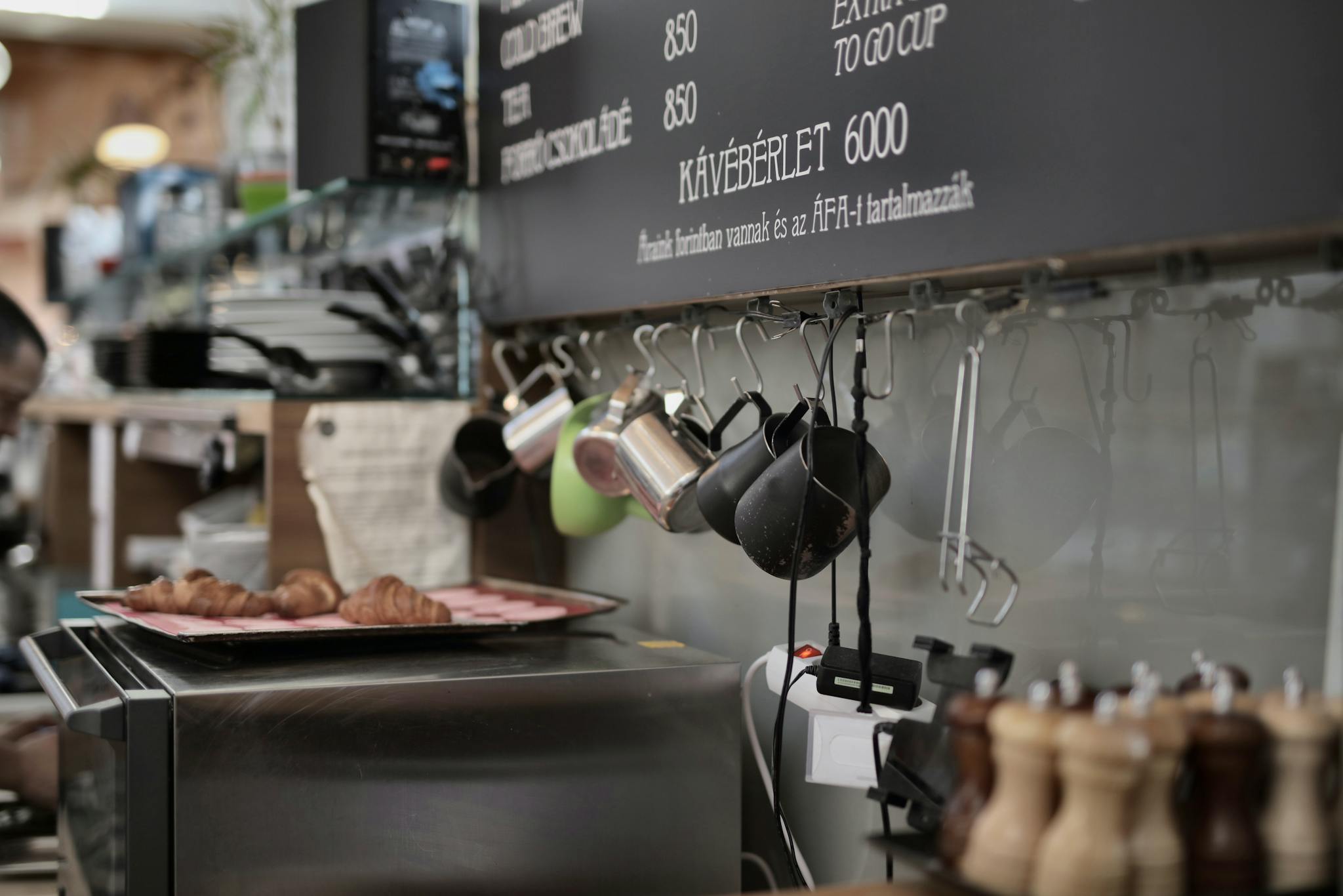 Various metal cups hanging on hooks near wall above oven with tray with freshly backed croissants in kitchen of modern cafe